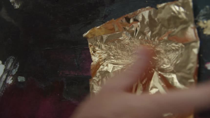 An artist placing gold foil onto a painted canvas and rubbing it in
