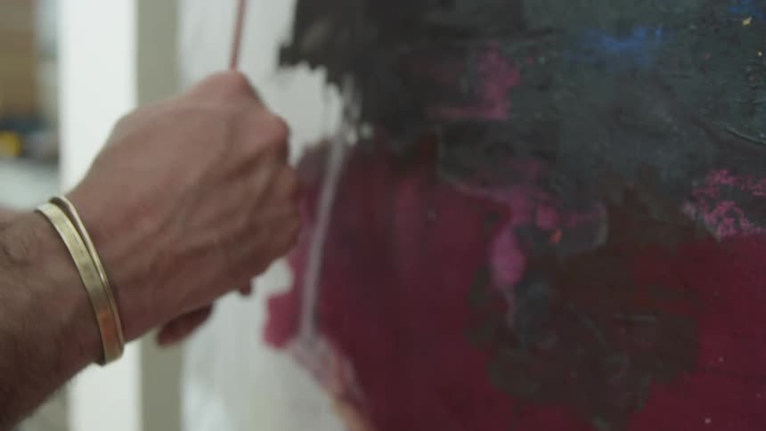 A man is holding a cup of paint in one hand and holding his paint brush with the other as he paints a picture on an easel