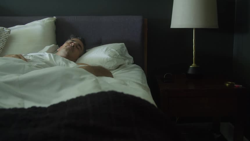 A man is sleeping in bed and there is a lamp on the nightstand next to the bed