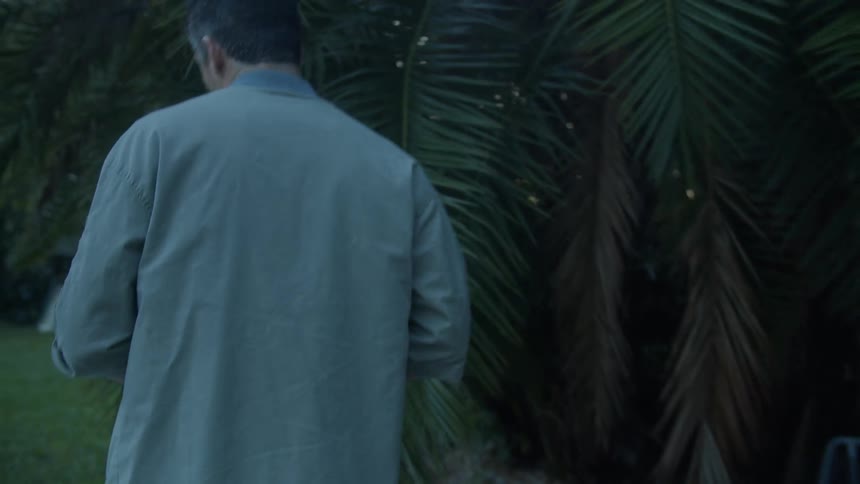 A man carrying a mug walks out of his house and past a swimming pool in to his back yard