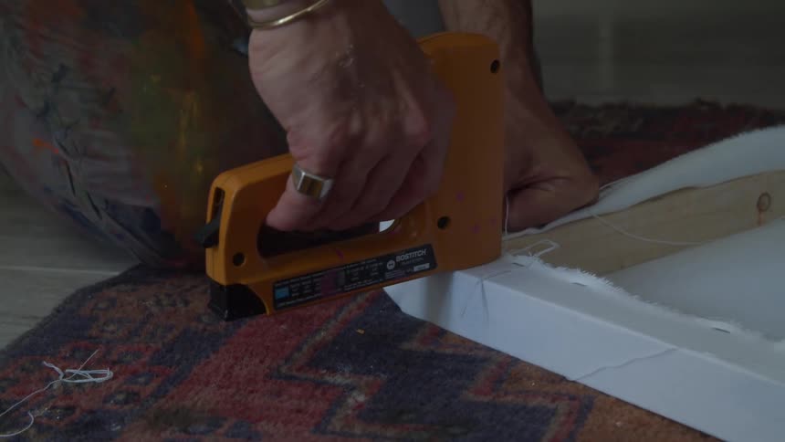 Kneeling on floor artist uses staple gun to attach fabric to wood to make canvas