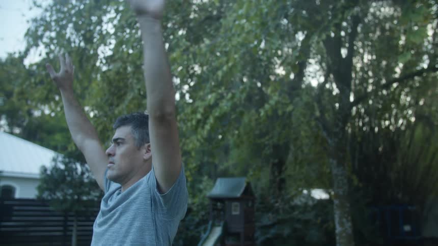 A man is standing outside in his yard exercising bending down and raising his arms over his head
