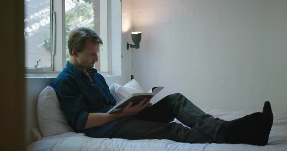 A man sits up in bed reading a book