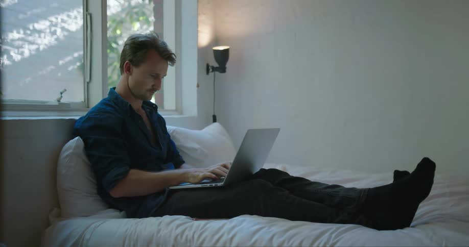 A man lays in bed and types on a laptop