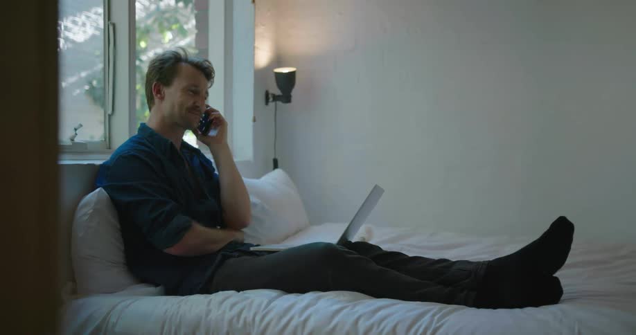 Man sits on a bed next to a laptop talking on a phone