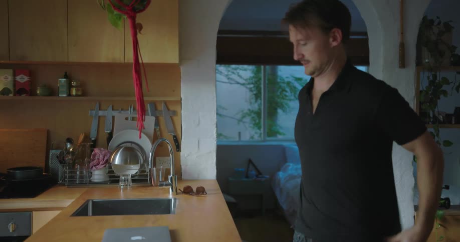 A man puts on a shirt while walking into a kitchen and opens a laptop