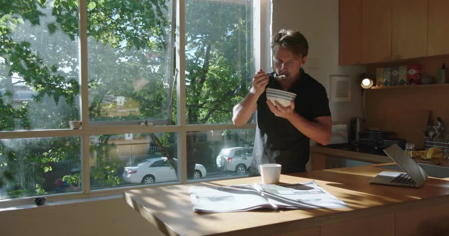 Man in kitchen reading newspaper while eating bowl of cereal during the day