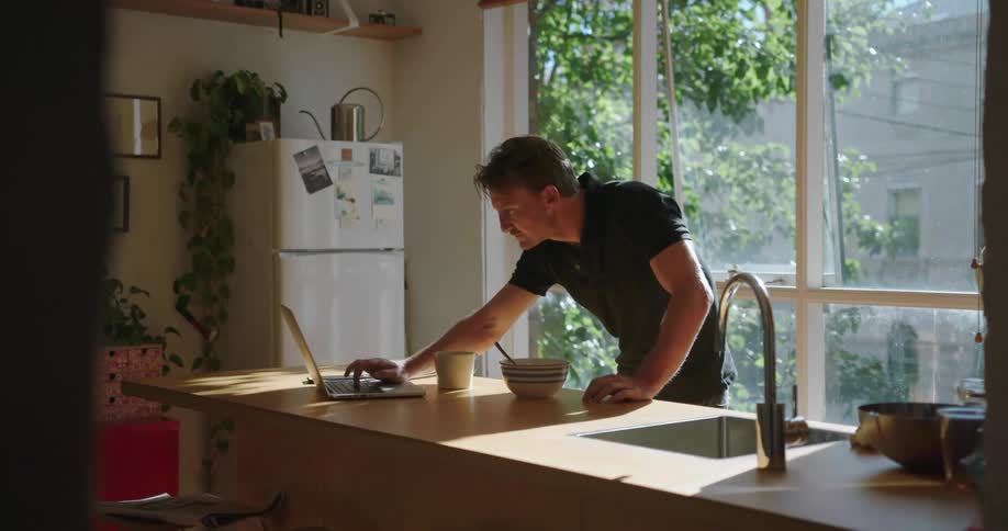 Man standing at a kitchen counter eating out of a bowl and typing on a laptop