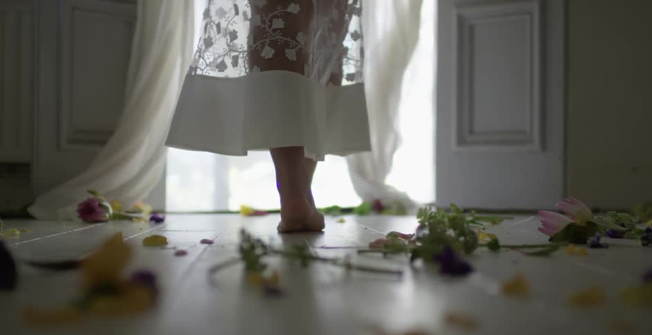 A barefoot woman crossing a floor strewn with flowers as she heads towards a doorway