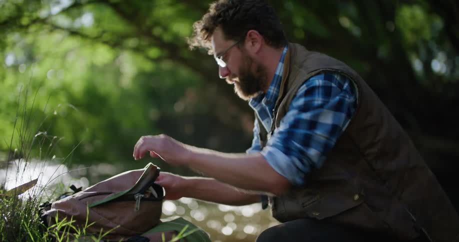 A man is standing in the woods near a stream and he places something in his backpack and lifts it over his shoulder