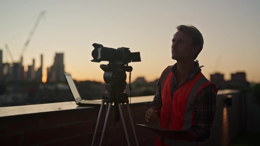 A man is standing behind a camera on a tripod and a laptop computer next to him overlooking the city as he writes on a clipboard