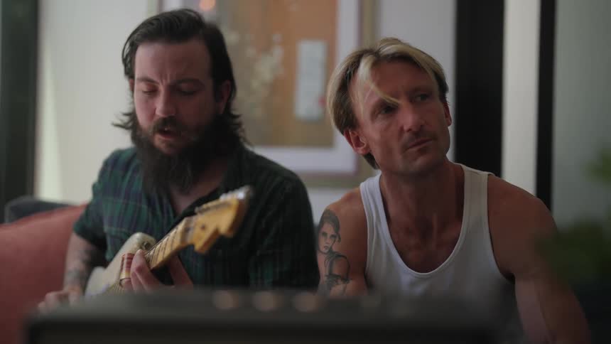 Two men sit on a couch while one sings and one thinks as he writes in a notebook