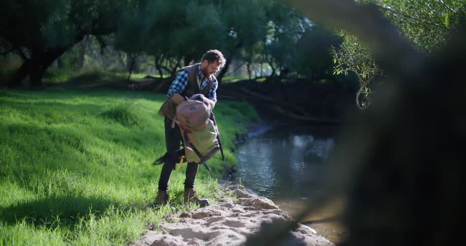 A man out hiking walks to a river and takes his back pack off and kneels down in front of the river
