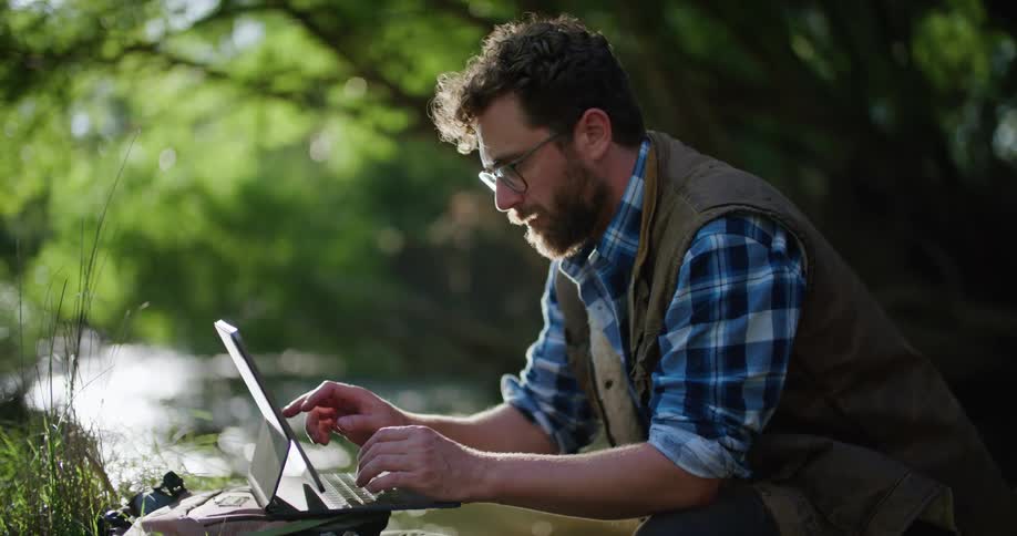 A man is kneeling in the woods near a stream as he types on his laptop computer