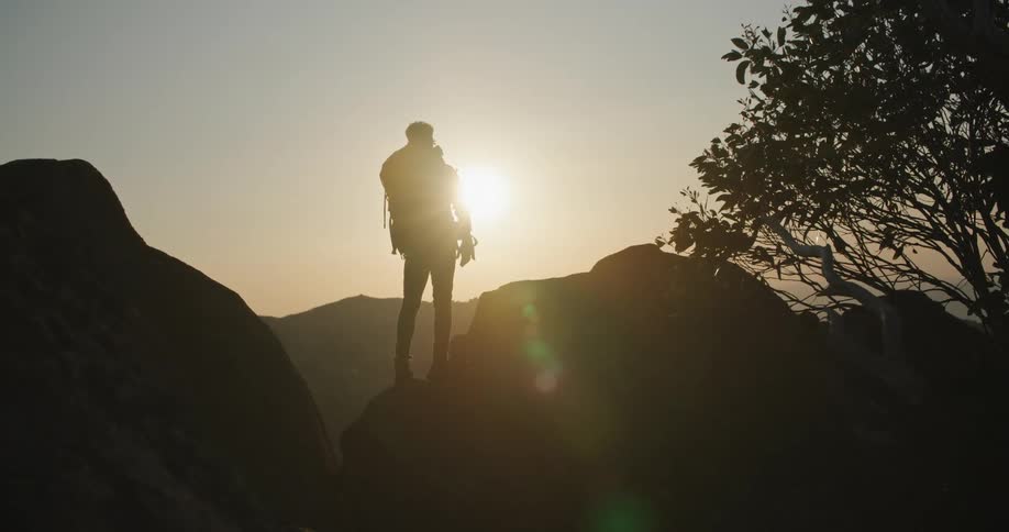 The sun shines brightly upon a man standing on rocks as he takes a picture of mountains in the distance