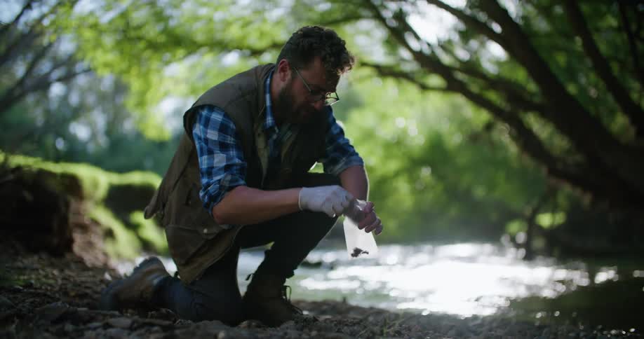 A man kneeling beside a river collects soil into a small plastic bag