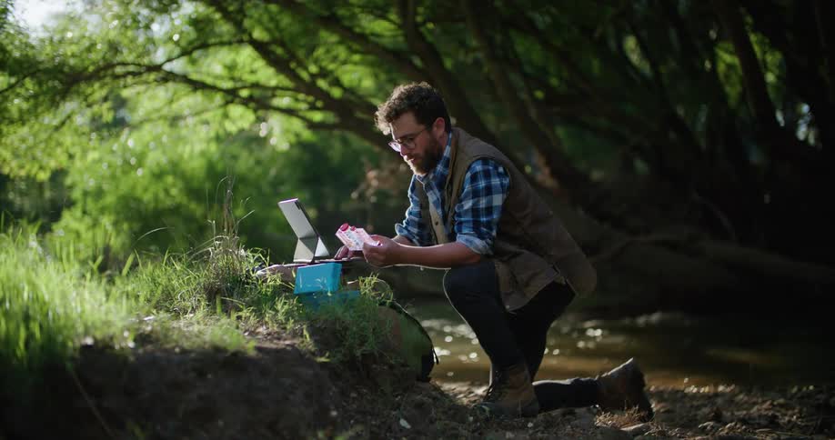 A man is kneeling in the woods near a stream as he holds a container in one hands and types on his laptop computer with the other hand
