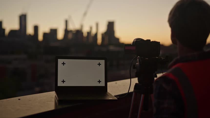 A man is standing behind a camera on a tripod and a laptop computer on a balcony overlooking the city