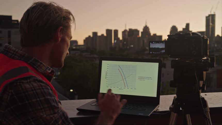 Man program the path of a drone in a laptop next to a camera in front of the city skyline at evening