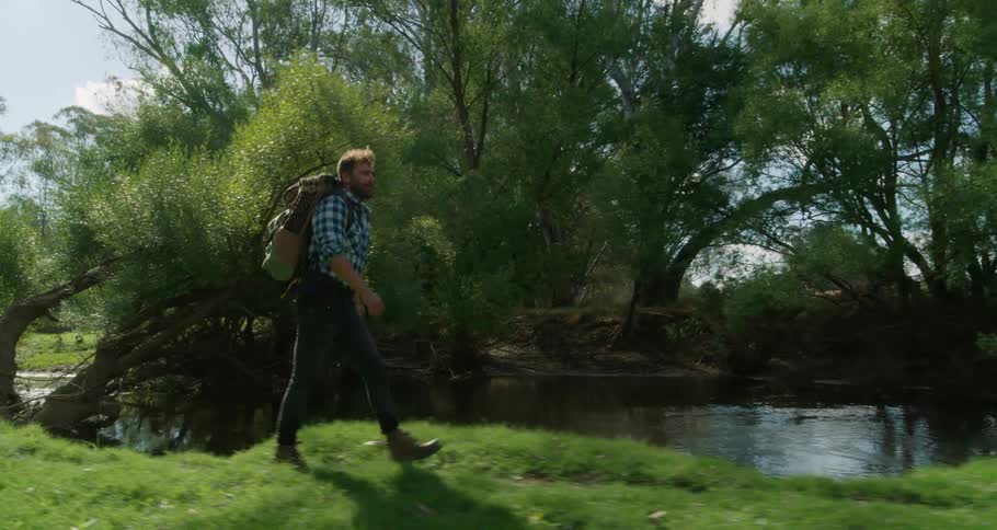 Male hiker walks along grass covered riverbank as bird flies past