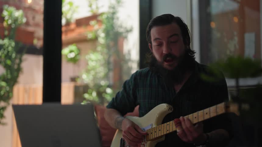 Man with beard and green plaid shirt plays guitar and sings a song while making a video call on laptop in room