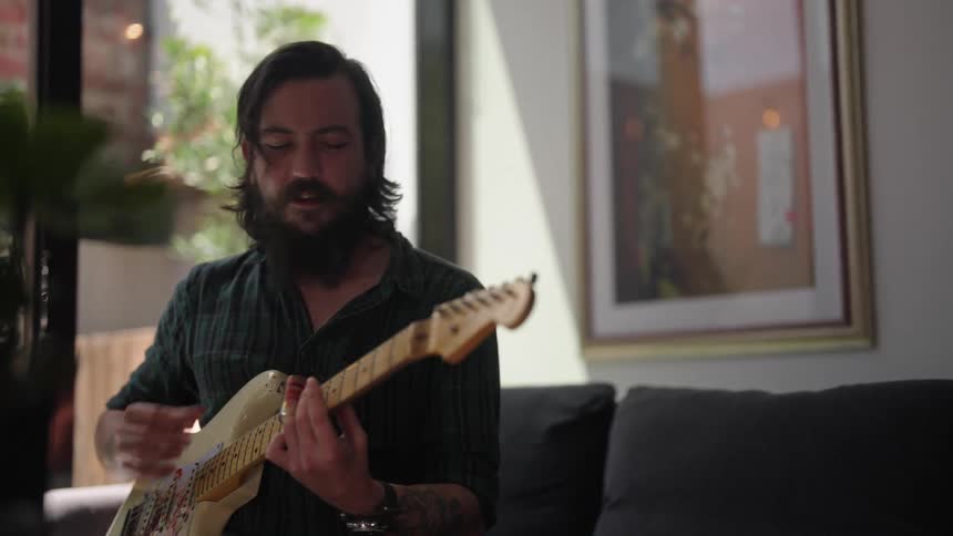 Man with beard and dark green shirt plays guitar and sings a song in room