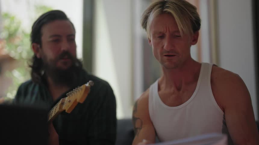 A man is sitting down holding a notebook and singing and a man next to him is playing a guitar