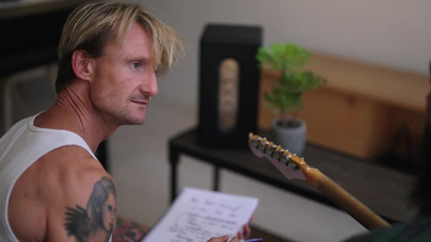 A man playing a guitar as another man writes lyrics in a notebook in a living room