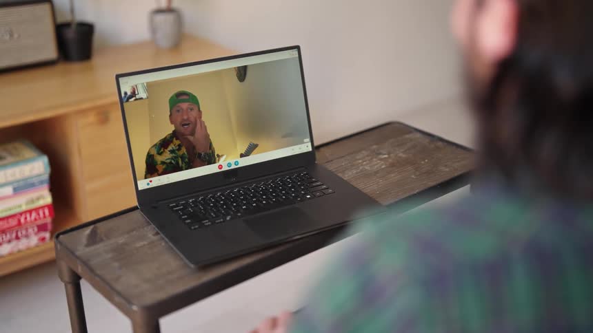 Man with green plaid shirt and beard makes a video call on laptop with man wearing green cap and shirt