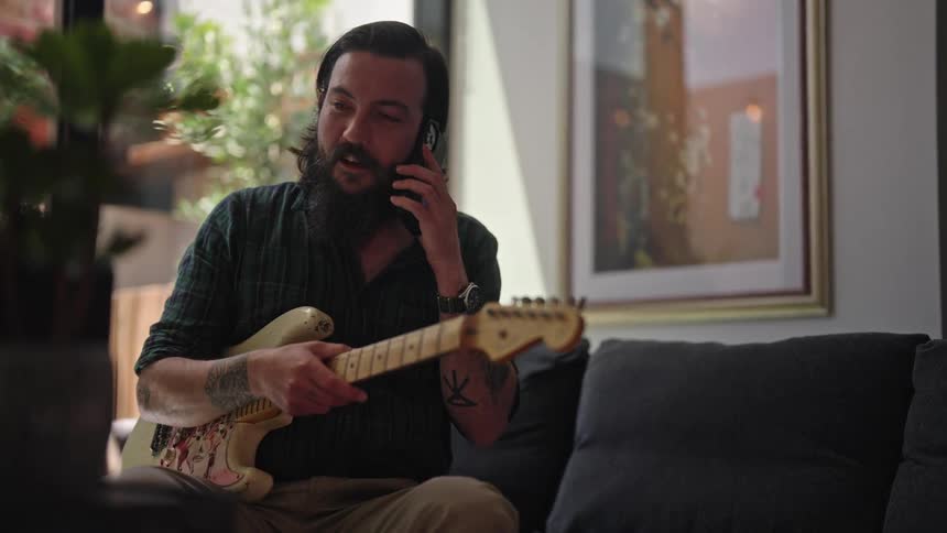 Man with beard green shirt brown pants and tattoos plays guitar and answers smartphone in room
