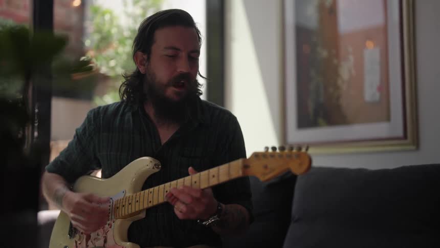 A man seated on a couch strums on his electric guitar while practicing a song