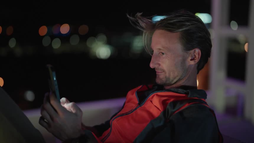 A man sits outside on his smartphone messaging someone with the night sky and city lights in the background