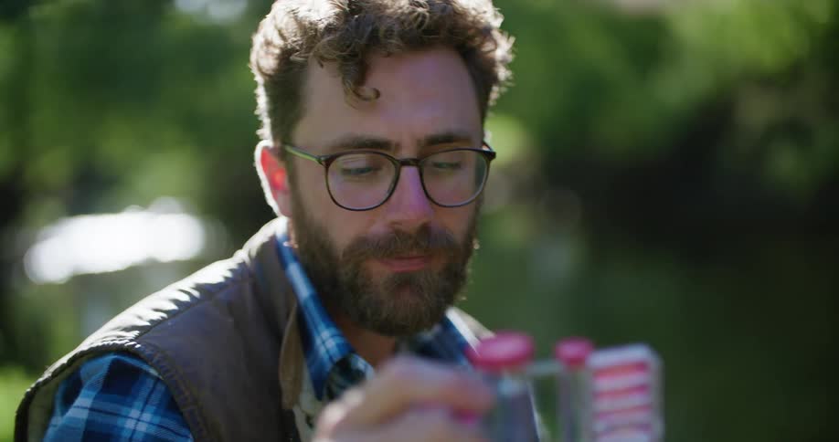 Man with beard glasses blue plaid shirt and vest holds test chamber near the stream
