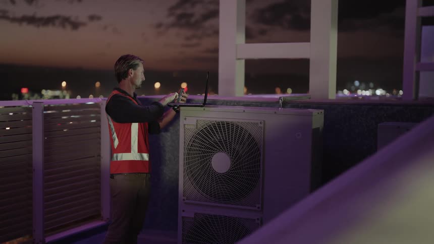 A male worker stands atop a building while evaluating some rooftop equipment