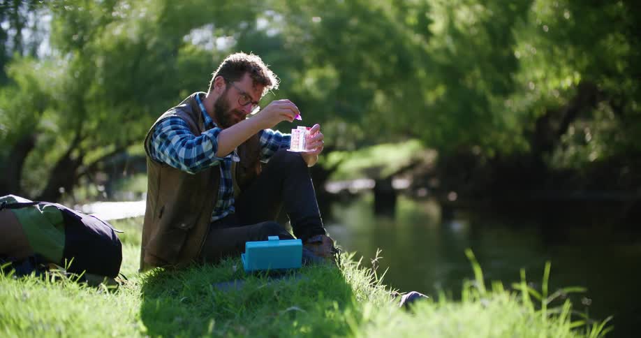 Man with glasses beard blue plaid shirt and brown vest uses test tubes near the river