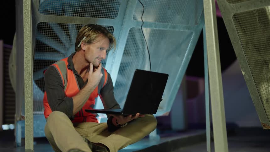 A man wearing a bright orange vest is sitting down and holding a laptop computer and typing then he closes it