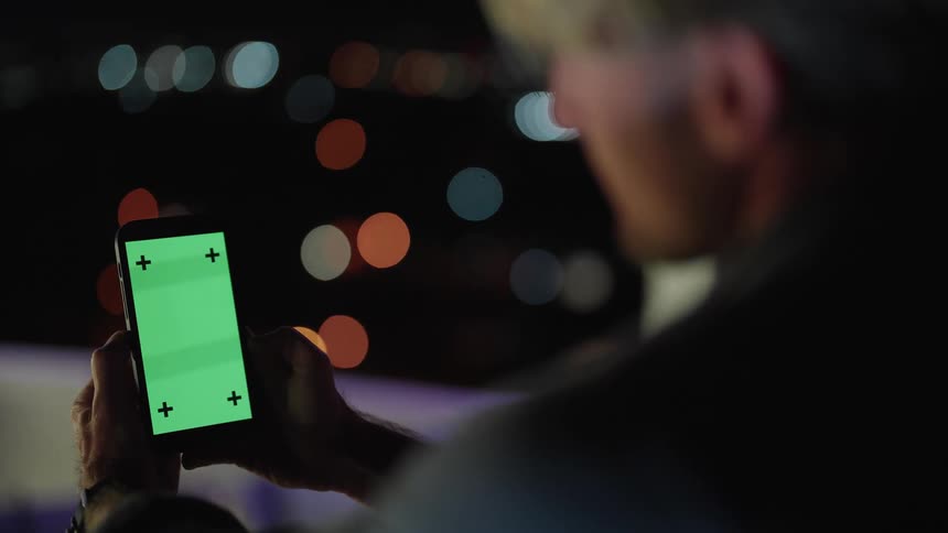 A person holds a phone with a green screen displayed with city lights flickering in the background