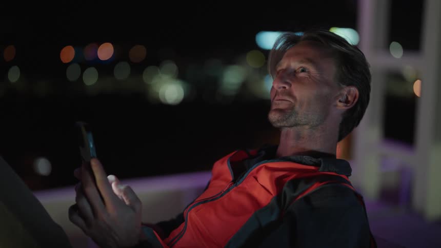 A man is sitting outside smiling and scrolling on his phone