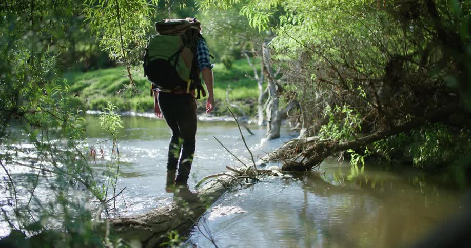 A man who is hiking walks across a river on a downed tree trunk