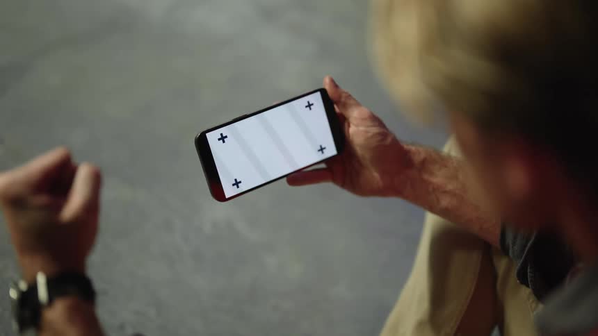 Blonde man wearing brown pants and gray shirt holds smartphone and clenches his fist