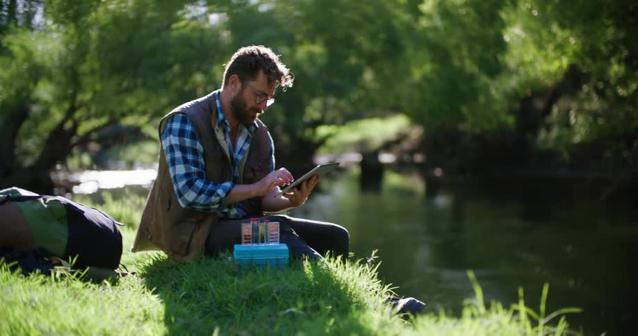 Male scientist sitting on riverbank enters data about water sample into tablet