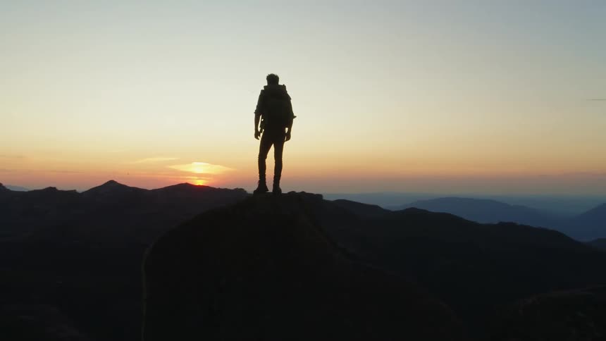 Man wearing backpack and standing on mountain is silhouetted against the sunset