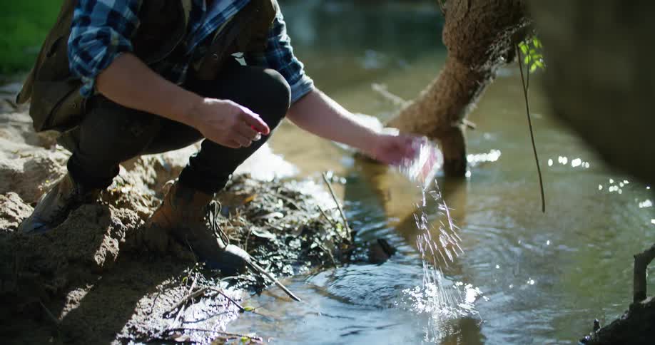 A man kneels in front of a river collecting some water in a measuring device and reading it