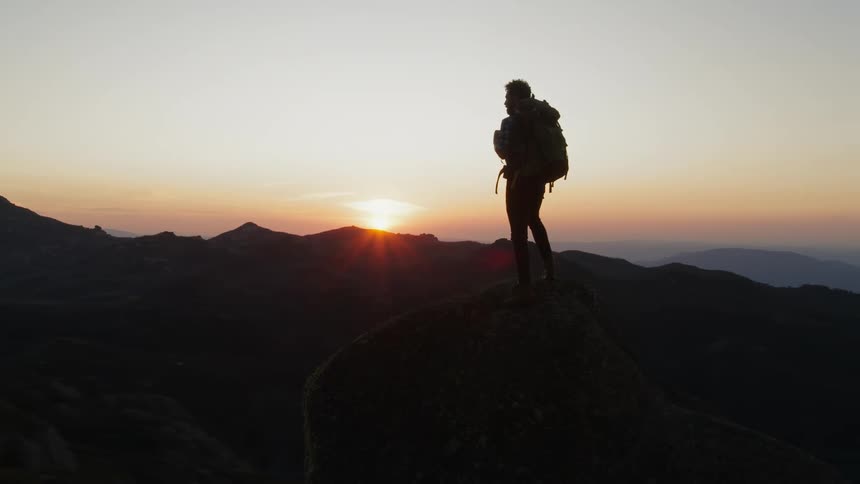 Man with backpack standing on mountain is silhouetted against the sunset