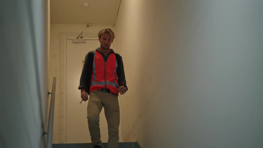 Middle aged man with safety vest backpack and brown pants opens door and goes downstairs in building