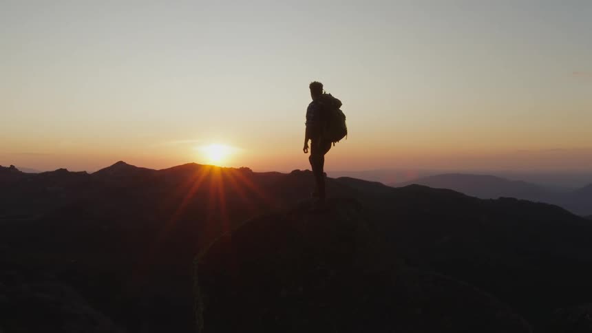 Man wearing backpack stands on mountain and is silhouetted against the sunset