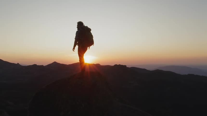Backpacker partially silhouetted by sun sitting on red horizon looks over foggy mountain range