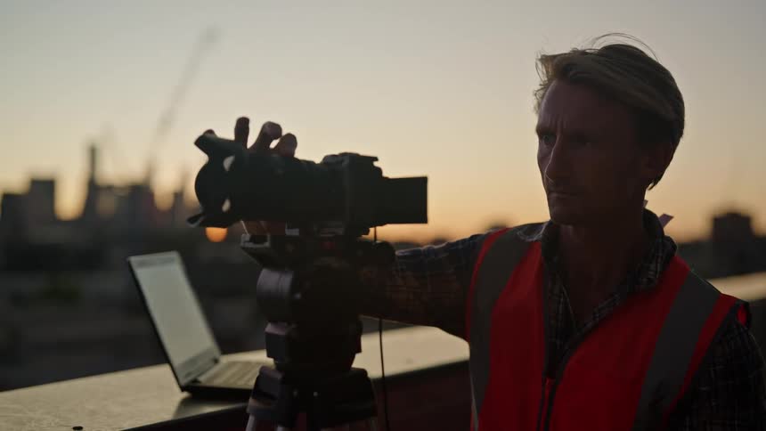 A man adjusting a video camera near a laptop outside