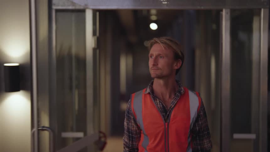 A man wearing a bright orange vest is walking down a hallway and out the door of a building