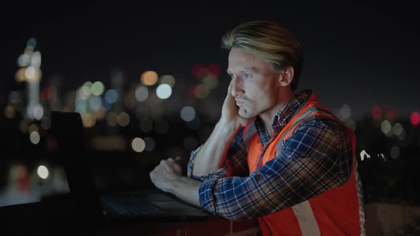 A man sits at his laptop typing outside at night with the city skyline lit up in the background